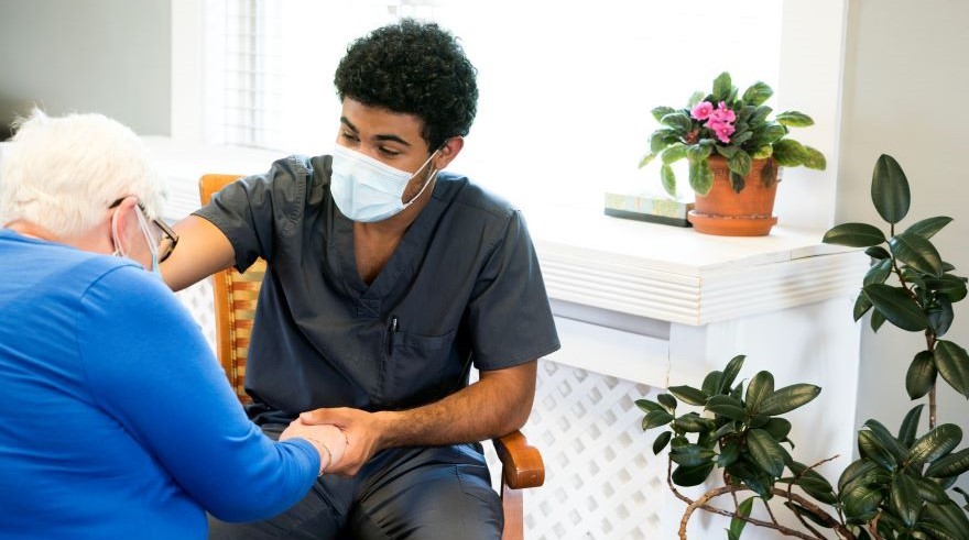 A male nurse with a mask on is helping an older female. 