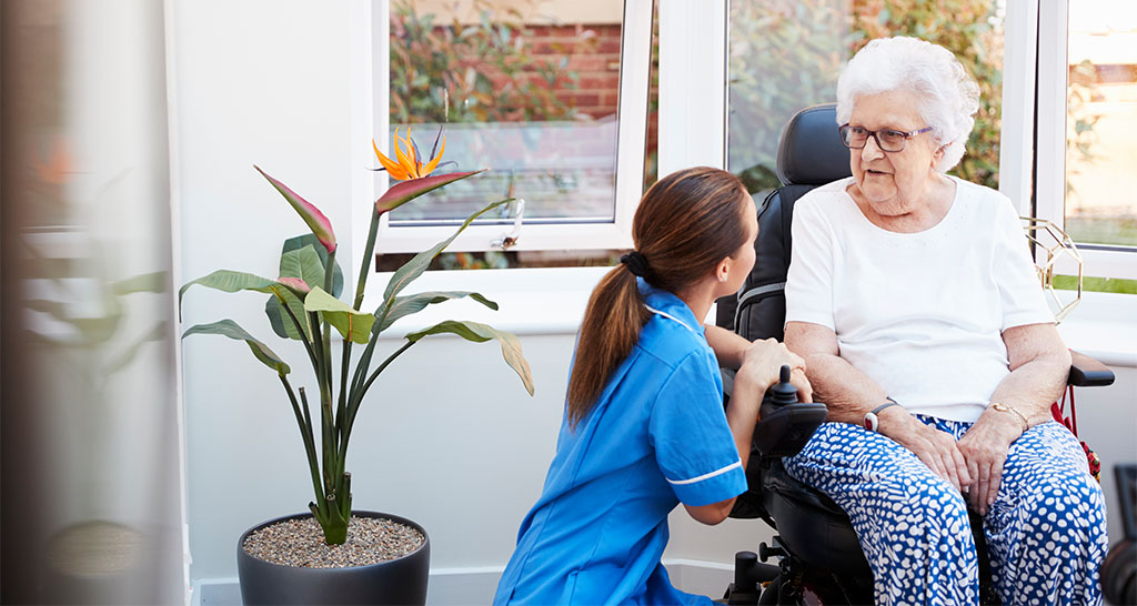 Senior Woman Sitting In Motorized Wheelchair Talking With Nurse In Retirement Home.