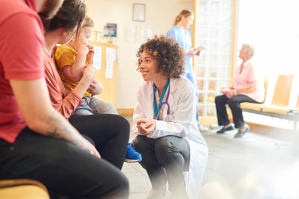 A doctor in white lab coat kneels and interacts with a toddler