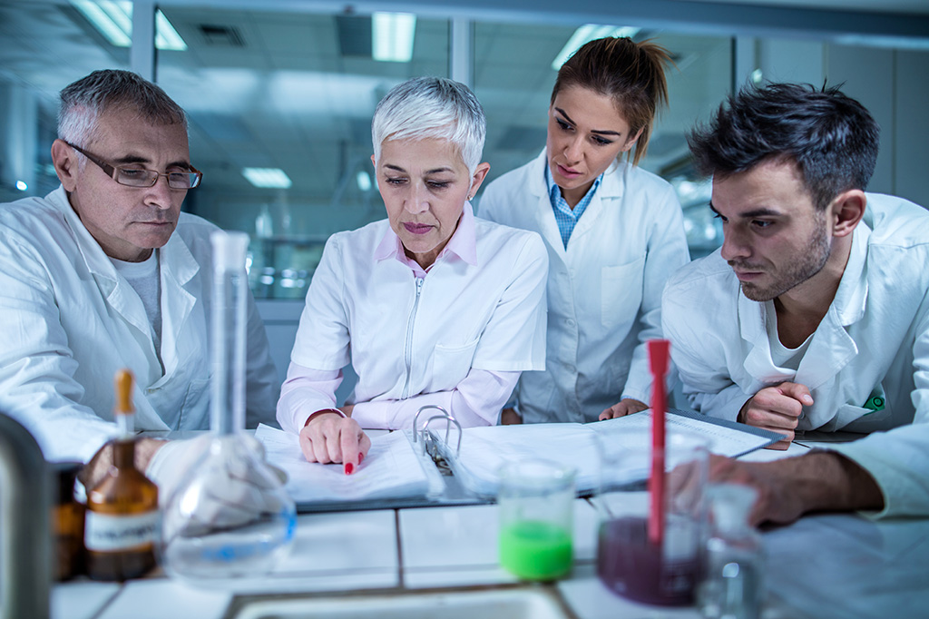 A group of doctors in white lab coats gather around a desk looking at a binder together.