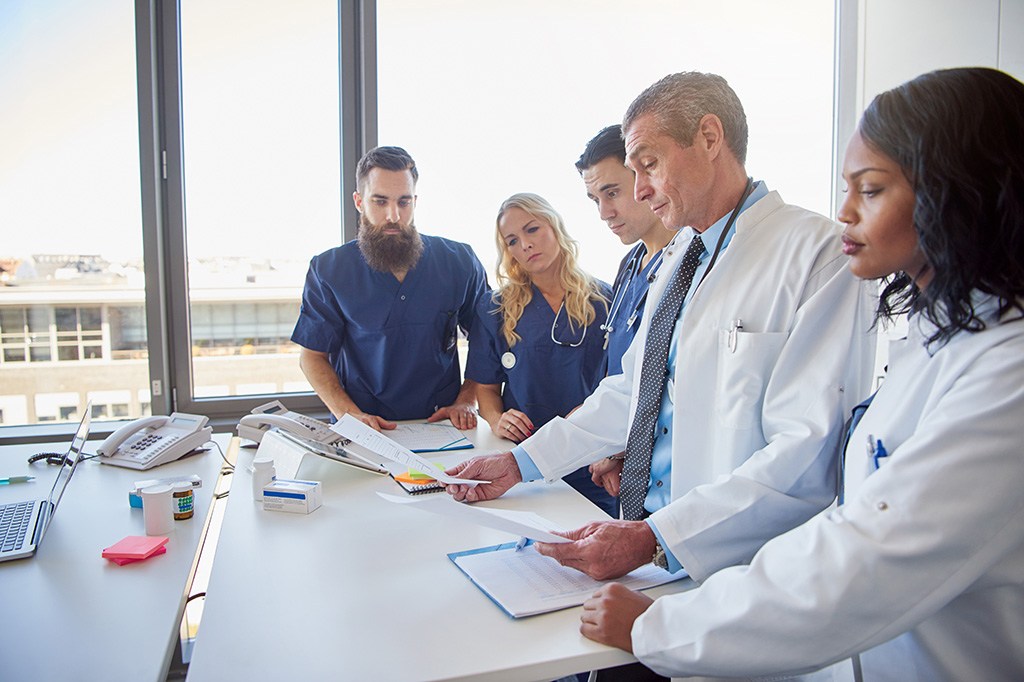 Two doctors in white lab coats and three nurses in blue scrubs gather around a table looking at documents.