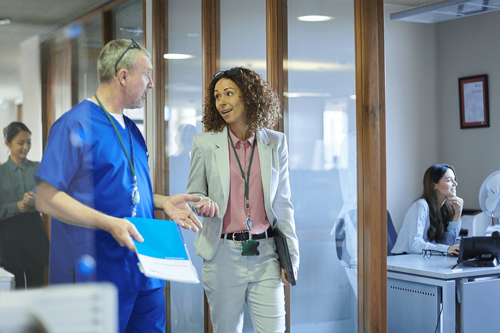 A male nurse in blue scrubs speaks to a woman in a business suit in a hospital hallway.