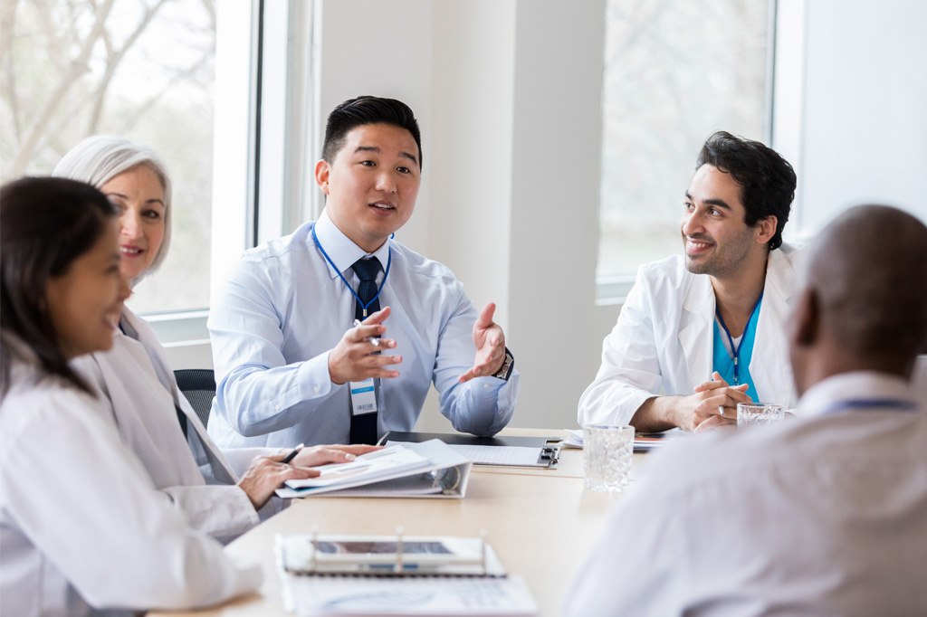 A surveyor speaks to a table of doctors in white lab coats.