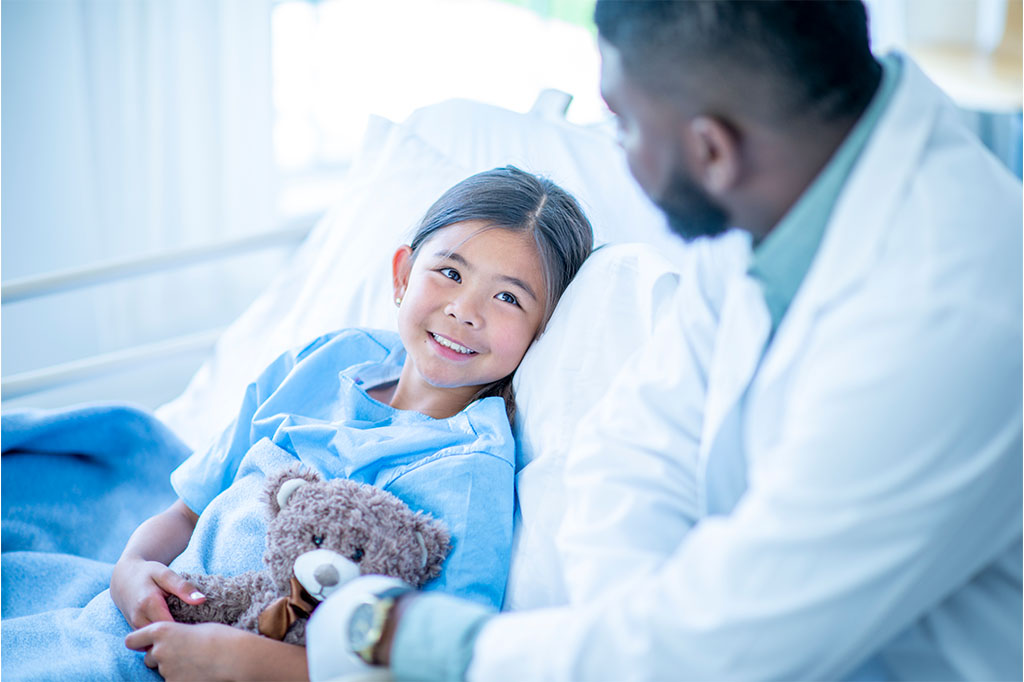 A doctor speaks with a young patient holding a stuffed animal in her hospital bed