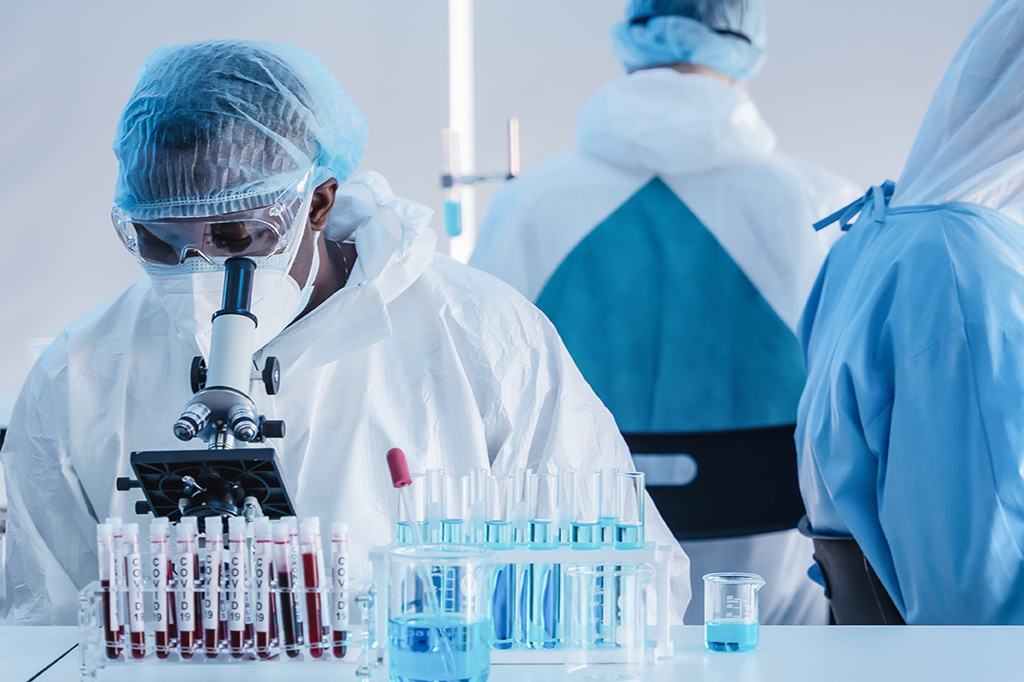 Three doctors work in a lab, looking into microscopes while wearing PPE. 